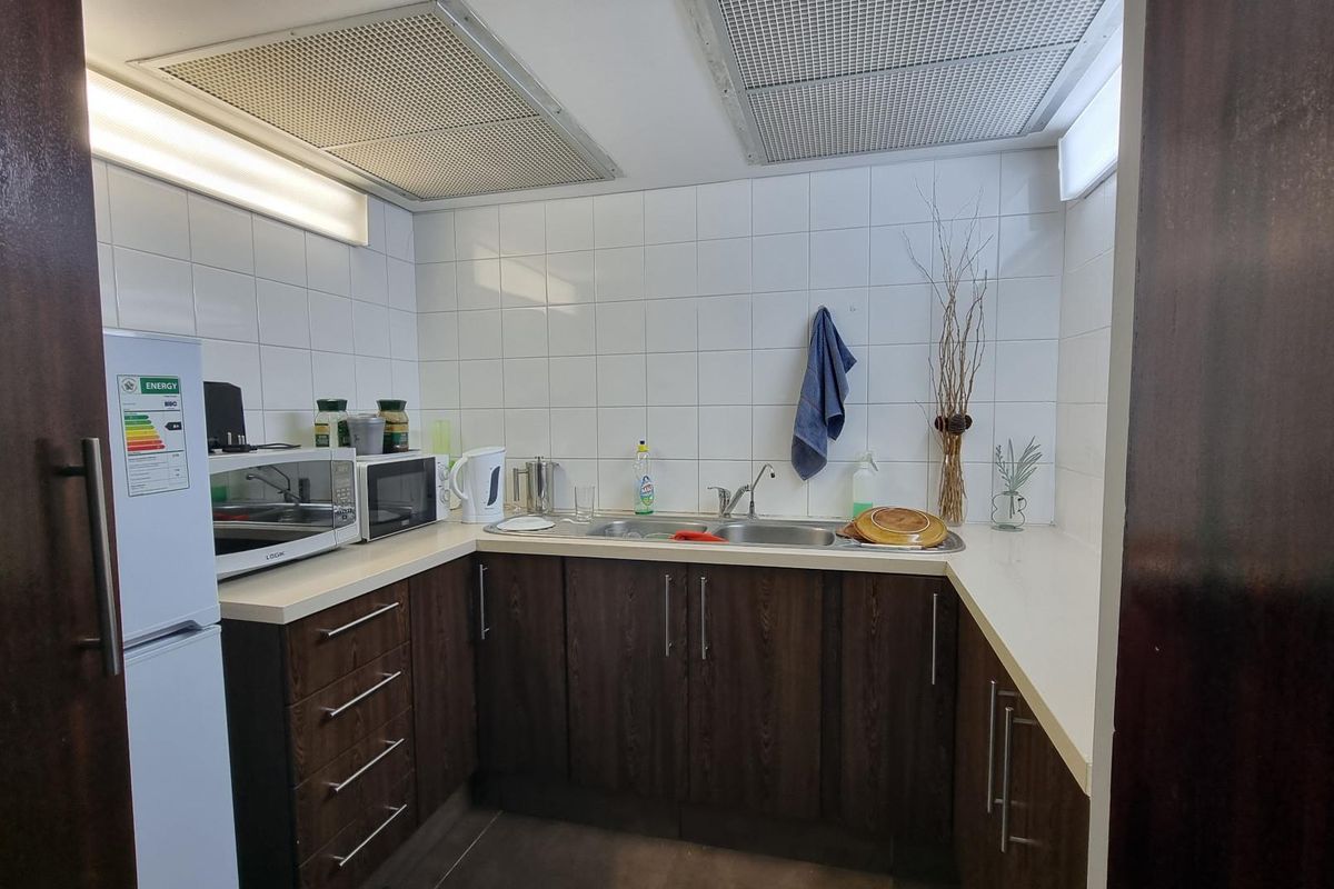 A compact office kitchenette featuring dark wood-finish cabinetry, a double-basin stainless steel sink, and white tiled backsplash with integrated ceiling ventilation.