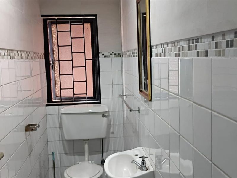 A compact, functional bathroom featuring white ceramic wall tiles, a pedestal basin, and a toilet with security bars on the window.