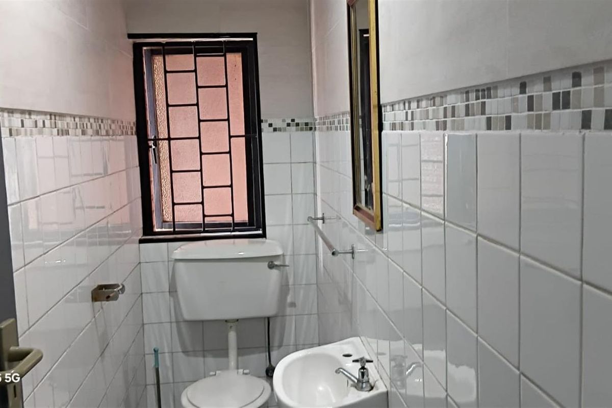 A compact, functional bathroom featuring white ceramic wall tiles, a pedestal basin, and a toilet with security bars on the window.