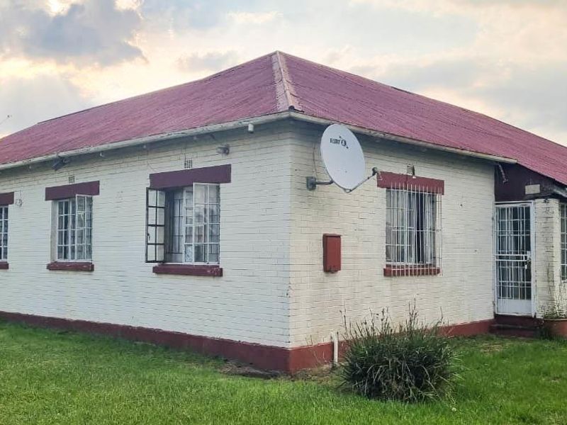 A detached single-story house featuring a corrugated metal roof, painted brick exterior, and security bars on the windows, set on a grassed plot.