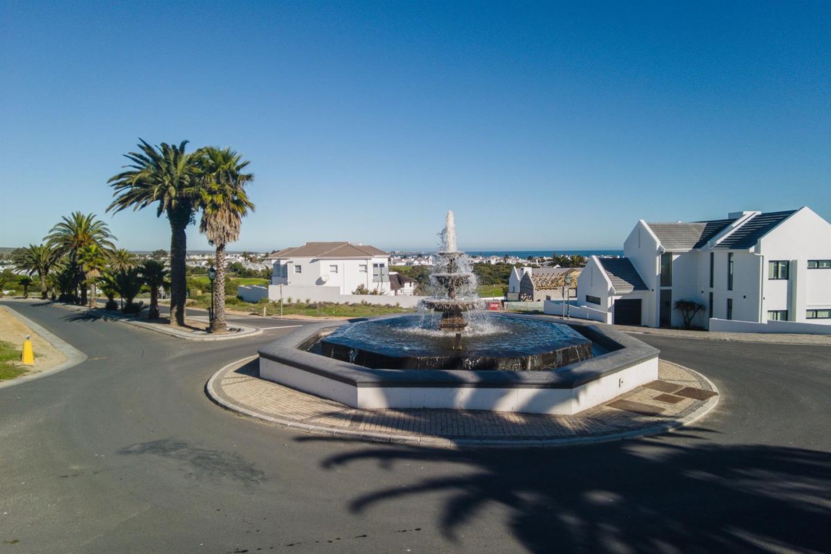 An aerial view of a residential estate entrance featuring a central tiered water fountain and paved roundabout surrounded by landscaped palm trees.