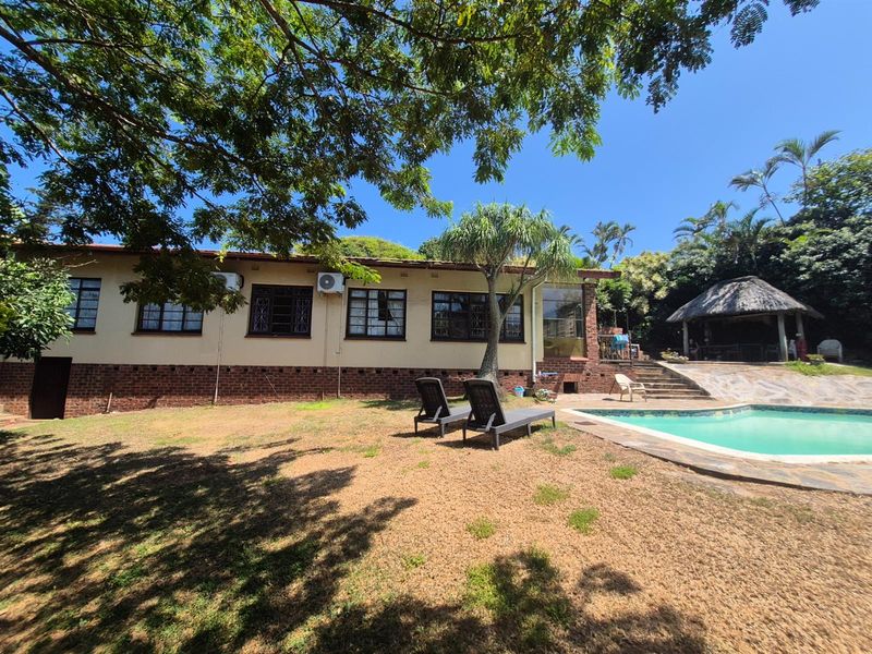 Rear view of the house featuring a swimming pool, paved pool area, and a thatched gazebo set within a spacious garden.
