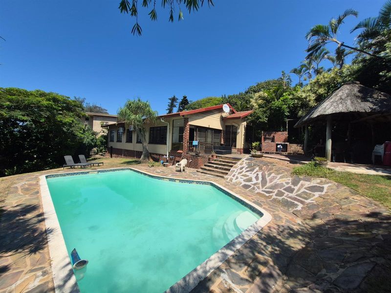 Rear view of the property featuring a swimming pool, stone paved patio area, and a thatched lapa structure set within a lush garden.