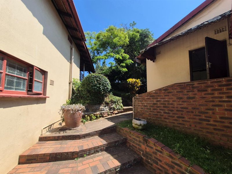 A paved outdoor walkway between two building structures featuring brick retaining walls and established garden landscaping.