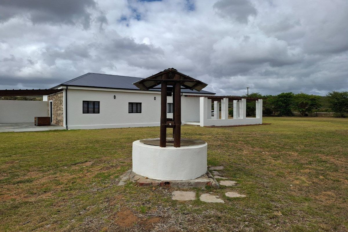 A view of the farm property exterior featuring a traditional stone-built water well in the foreground and a modern white-walled dwelling with pergola structures.