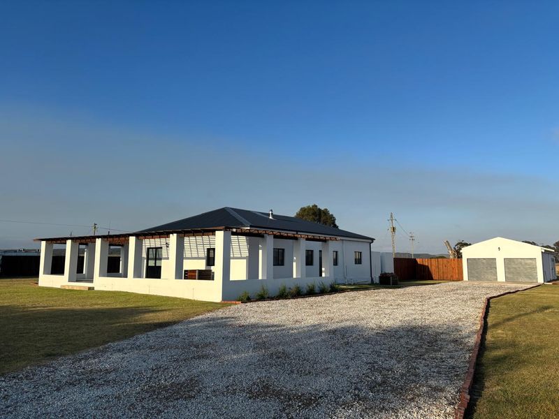 A well-maintained farm property featuring a white exterior, a covered wrap-around patio, a gravel driveway, and a detached double garage.
