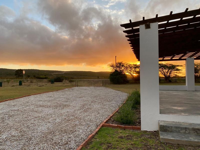 A scenic view of a farm property at sunset, featuring a covered patio area with a pergola and a gravel driveway.