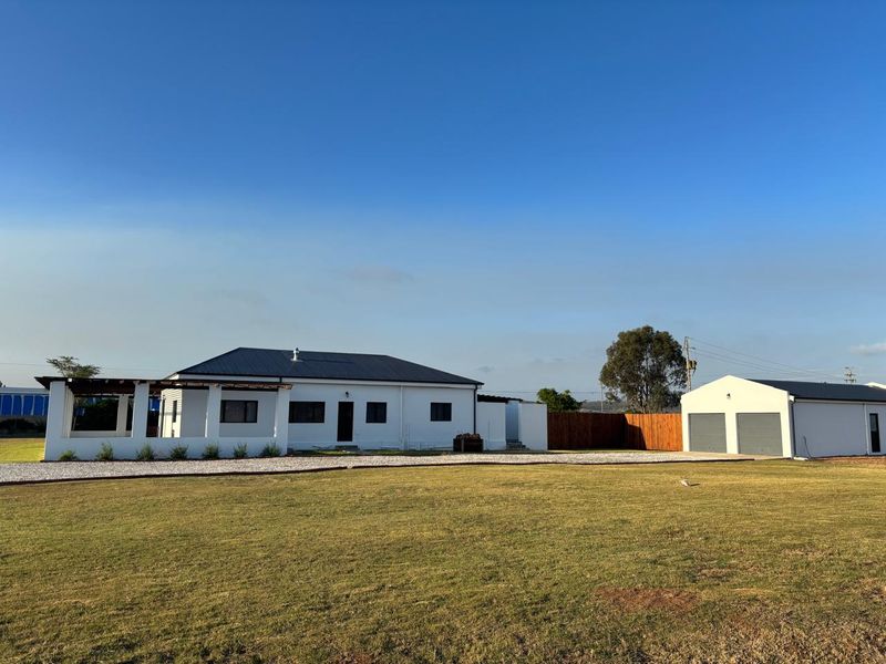 A well-maintained farm property featuring a white exterior, a covered patio area, and a separate detached double garage set on a spacious lawn.