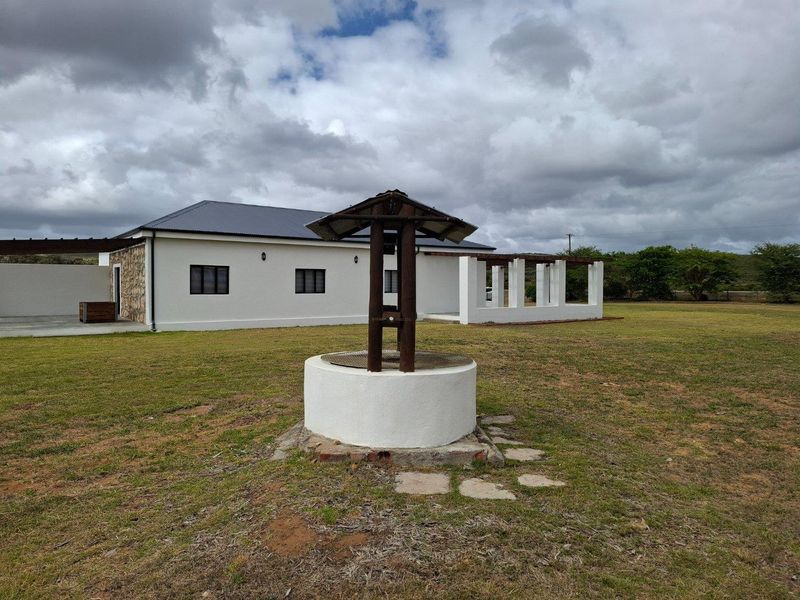 A view of the farm property exterior featuring a traditional stone-built water well in the foreground and a modern white-walled dwelling with pergola structures.