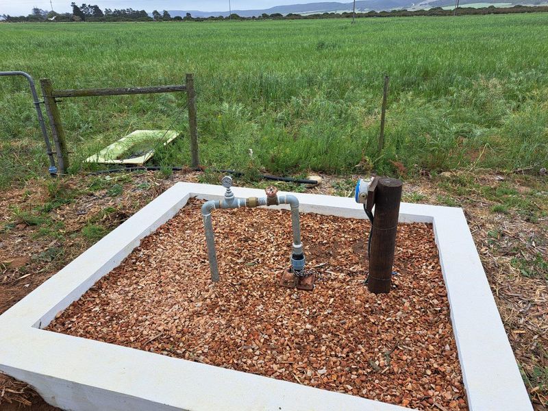 A functional borehole pump installation set within a concrete-bordered gravel enclosure, situated in an open agricultural field.