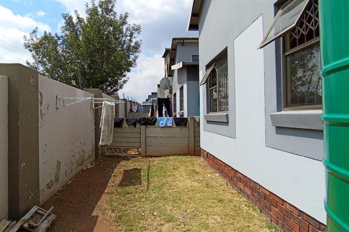 A side exterior view of the property featuring a small lawn, boundary wall with security fencing, and a water storage tank.
