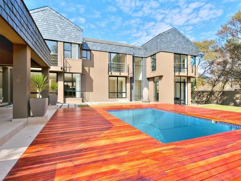 A modern rear exterior view featuring a swimming pool with a timber deck, paved patio area, and multi-level architectural facade with large glass windows.