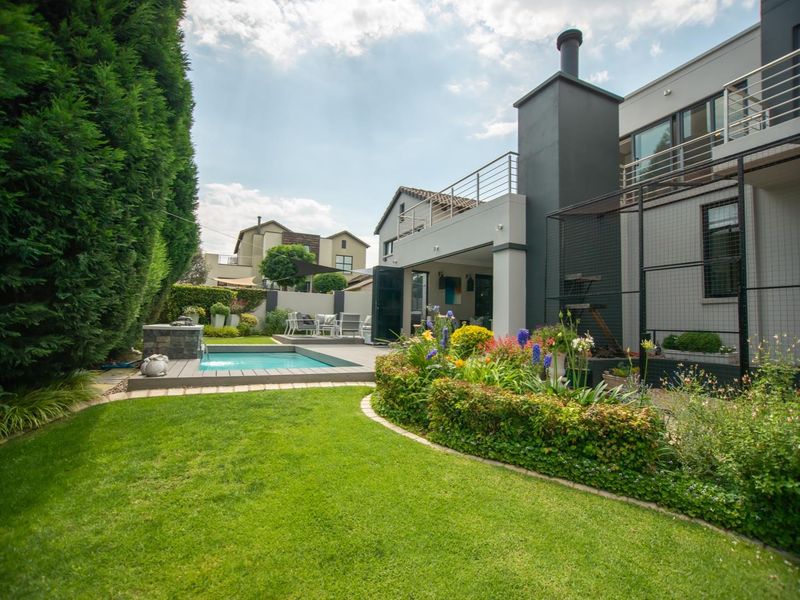 A well-maintained rear garden featuring a swimming pool, wooden decking, an outdoor fireplace, and a balcony overlooking the landscaped yard.