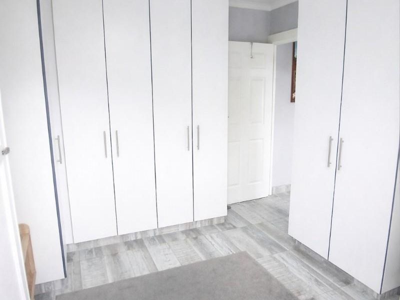 A clean, bright bedroom featuring extensive white built-in cupboards, modern wood-look laminate flooring, and a contemporary flush-mount ceiling light fixture.