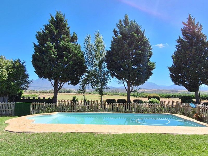 Outdoor swimming pool area set within a landscaped garden, featuring a paved surround, perimeter fencing, and scenic mountain views.