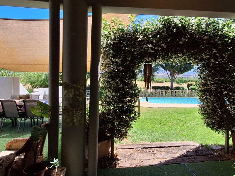 Covered patio area overlooking a lush garden, swimming pool, and scenic landscape, featuring a shaded outdoor dining space and a decorative floral archway.