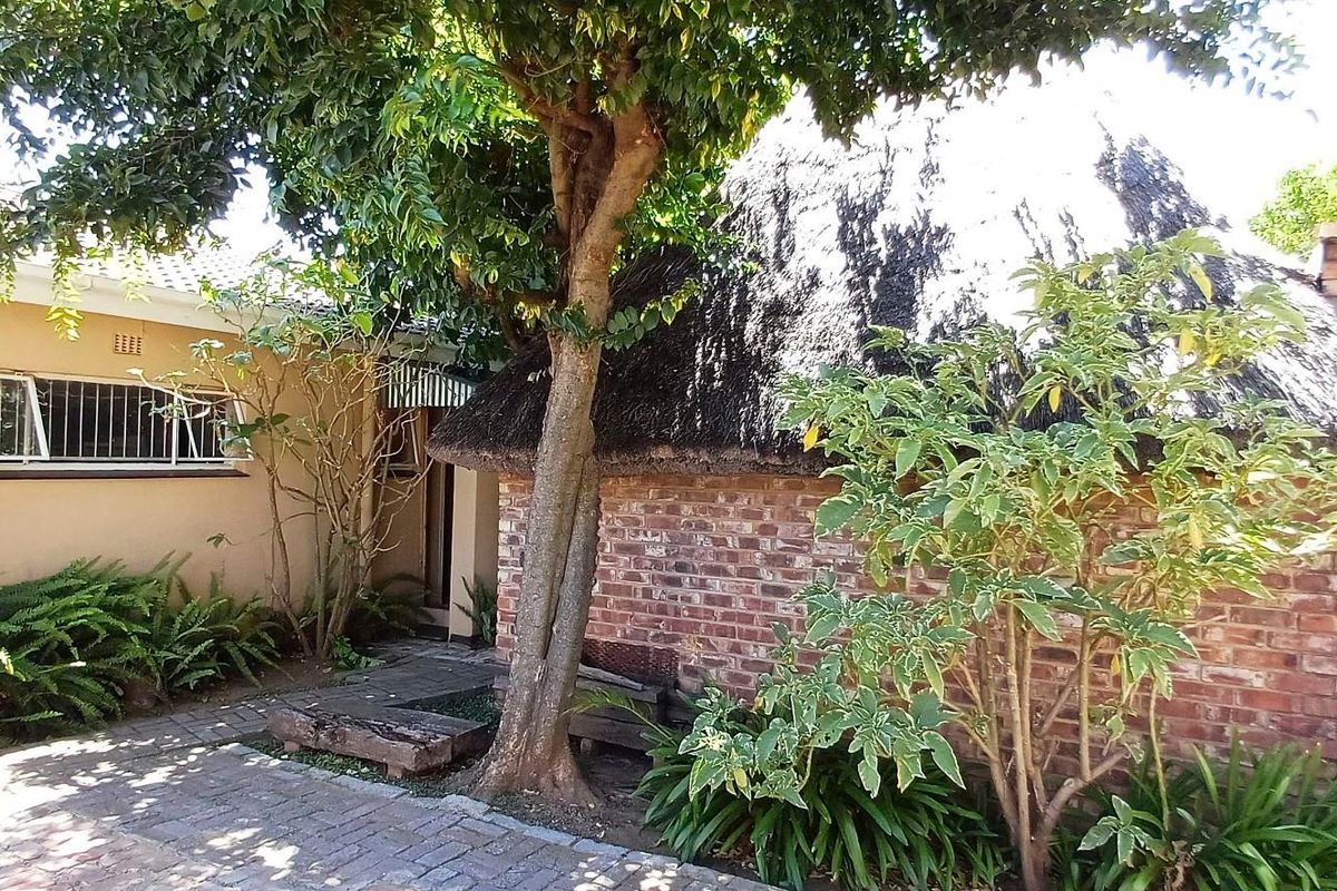 Rear exterior view featuring a thatched roof structure, brick fireplace, and paved patio area surrounded by established garden greenery.