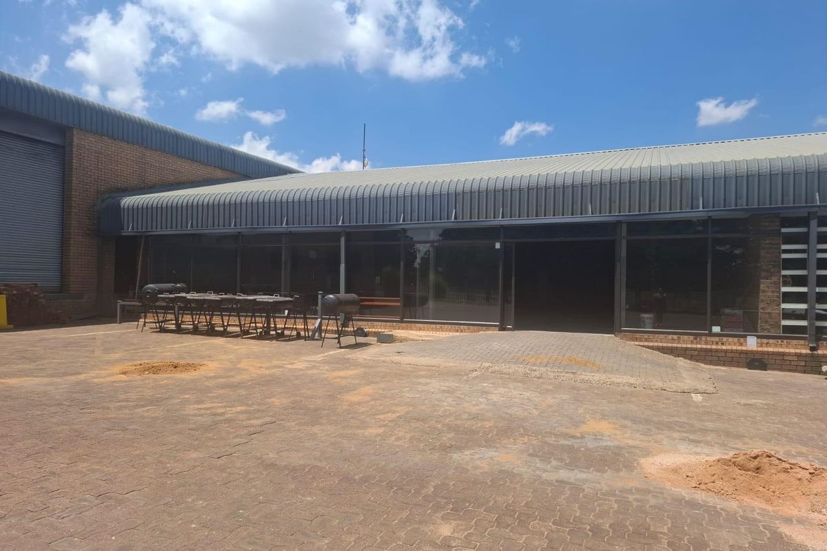 Exterior view of a retail property featuring a large paved courtyard, expansive glass frontage, and a covered walkway with metal roofing.