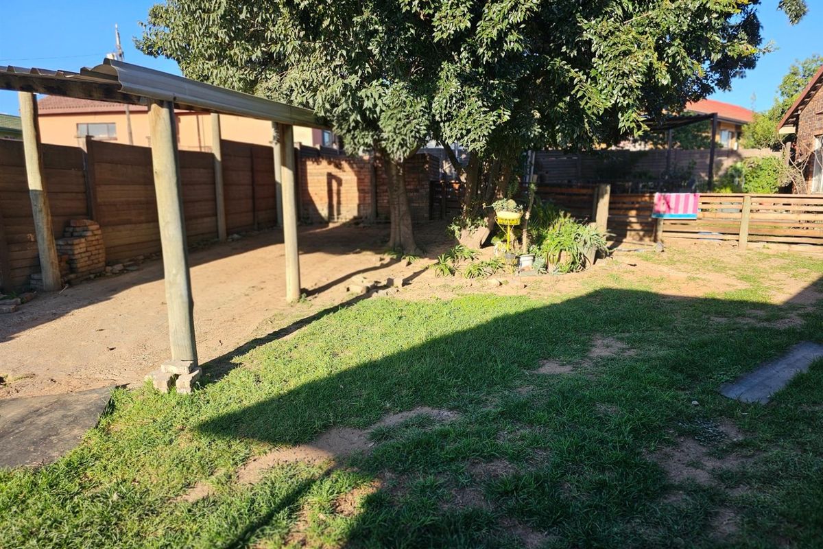 A spacious rear garden area featuring a lawn, mature trees, and a simple covered outdoor structure with timber fencing.