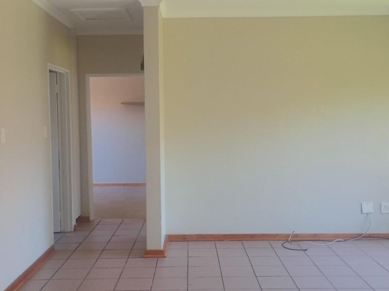 An empty living area featuring neutral walls, tiled flooring, and a window with security bars, leading into an adjacent room.