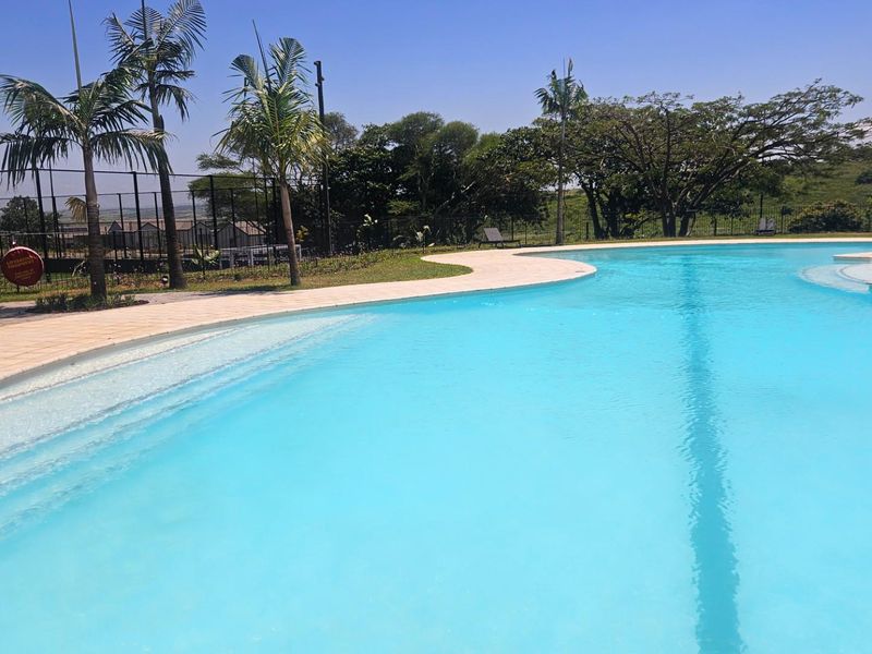 A well-maintained communal swimming pool area featuring a paved surround, perimeter fencing, and established landscaping in a residential estate setting.