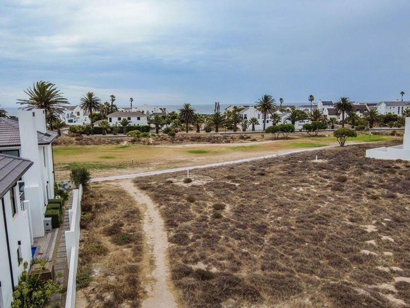 Aerial view of a vacant residential plot featuring natural vegetation, a paved pathway, and adjacent modern housing with solar panels.