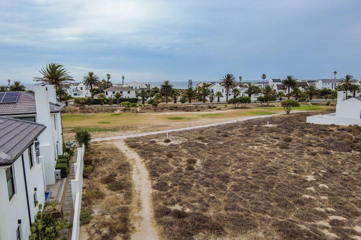 Aerial view of a vacant residential plot featuring natural vegetation, a paved pathway, and adjacent modern housing with solar panels.