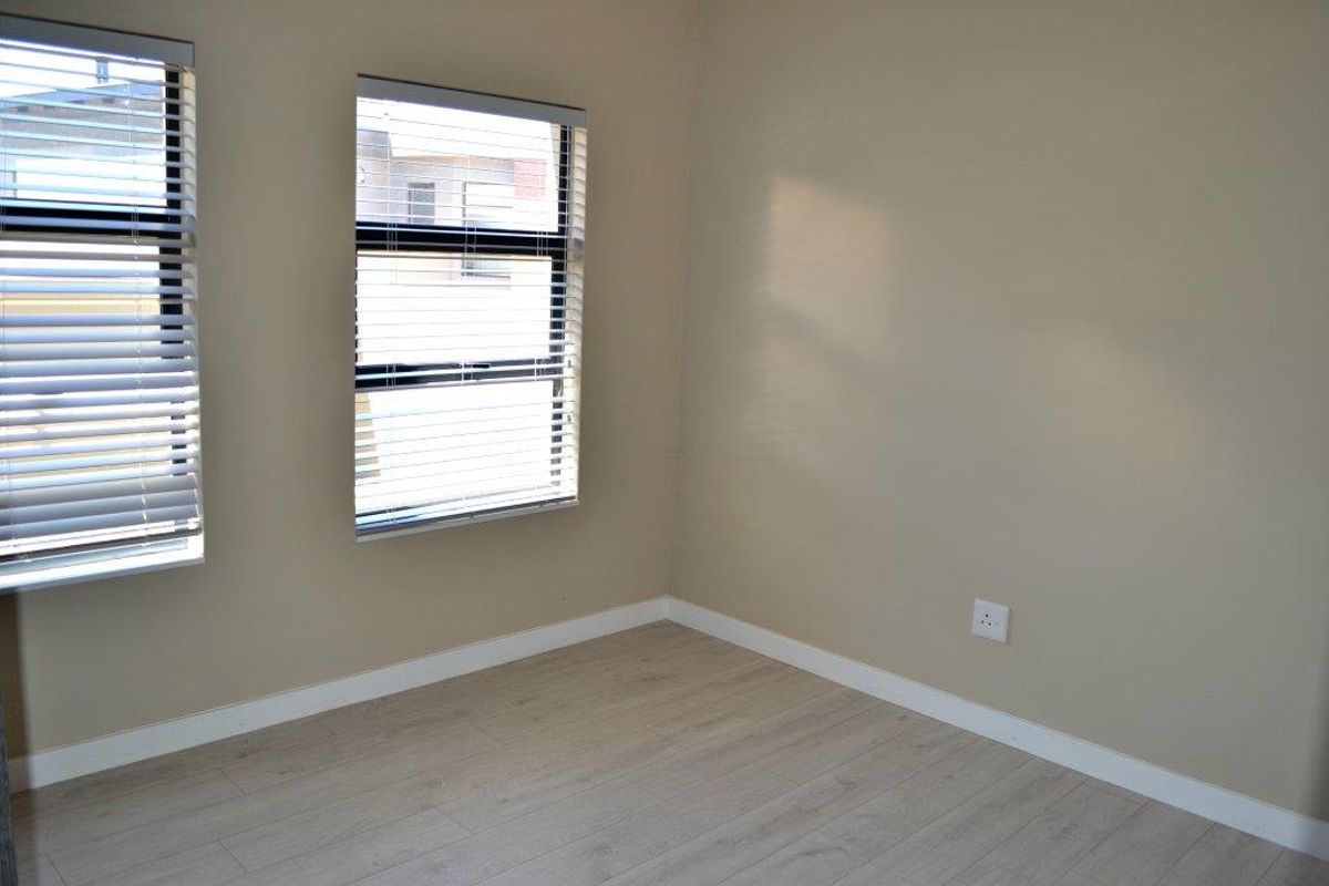 A bright, empty bedroom featuring light-toned laminate flooring, neutral wall paint, and fitted venetian blinds on the windows.