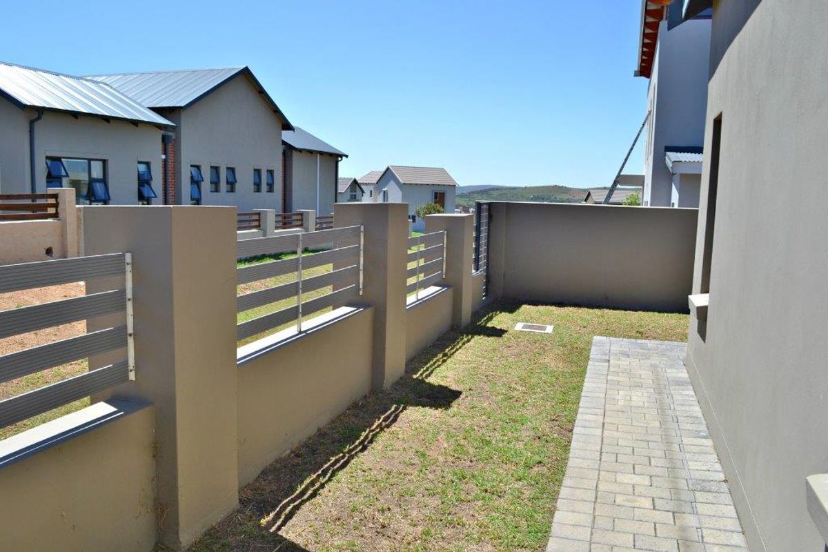 A side garden area featuring a paved walkway, manicured lawn, and modern boundary wall with metal slat fencing.