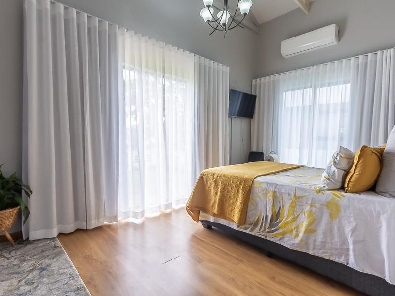 A bright and airy bedroom featuring laminate flooring, a vaulted ceiling, split-system air conditioning, and large sliding glass doors with floor-to-ceiling curtains.