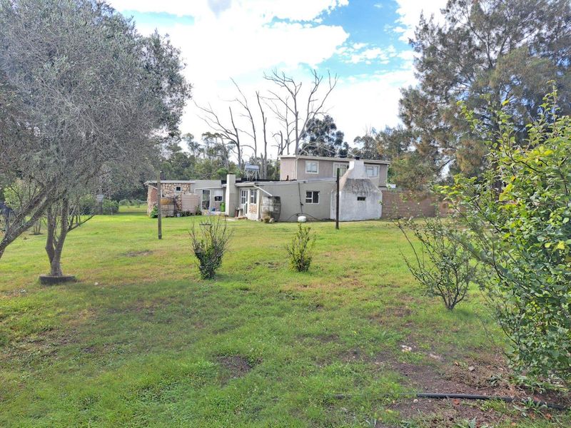 Rear view of a farm property featuring a large garden, outdoor fireplace, and established trees, showcasing the rural landscape and building exterior.