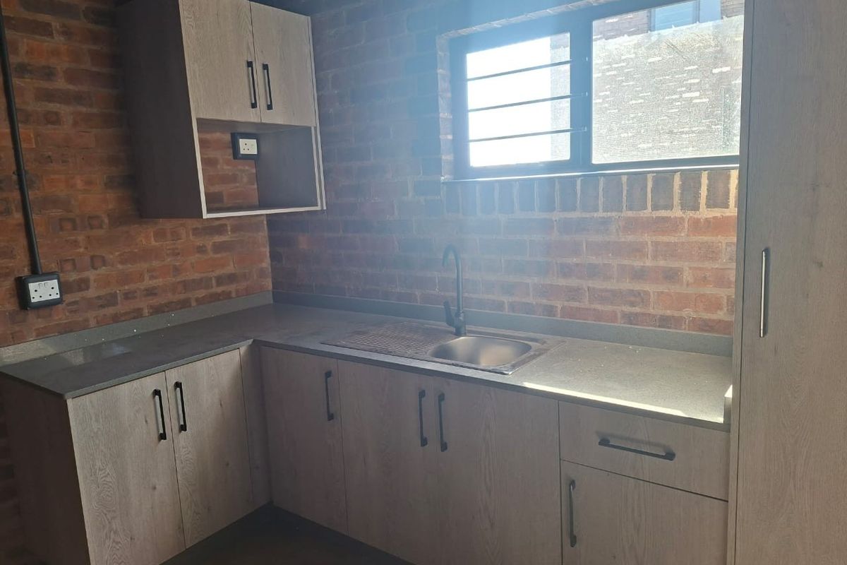 A compact kitchen featuring exposed brick walls, modern laminate cabinetry, stone countertops, and a stainless steel sink with a contemporary black mixer tap.