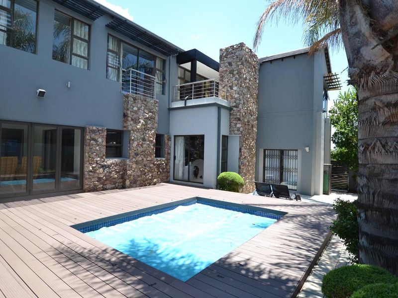 A well-maintained rear exterior featuring a swimming pool with a deck, stone-clad architectural details, and a balcony overlooking the outdoor entertainment area.