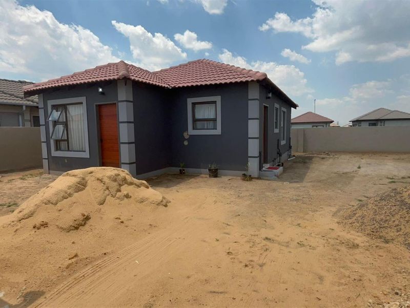 Exterior view of a detached house featuring a dark grey facade with tiled roof and perimeter boundary walls, set on an undeveloped plot.