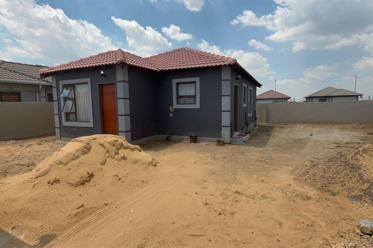 Exterior view of a detached house featuring a dark grey facade with tiled roof and perimeter boundary walls, set on an undeveloped plot.