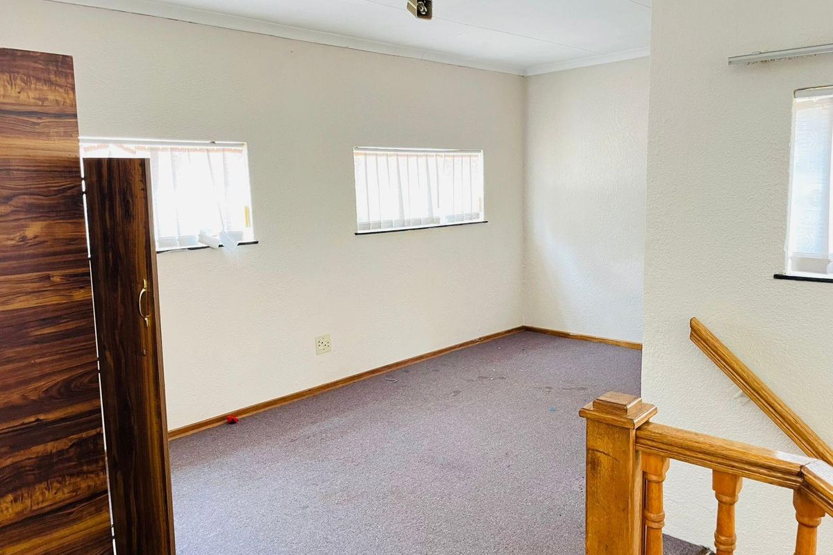 An empty bedroom featuring carpeted flooring, two windows with vertical blinds, and a wooden staircase railing leading to the space.