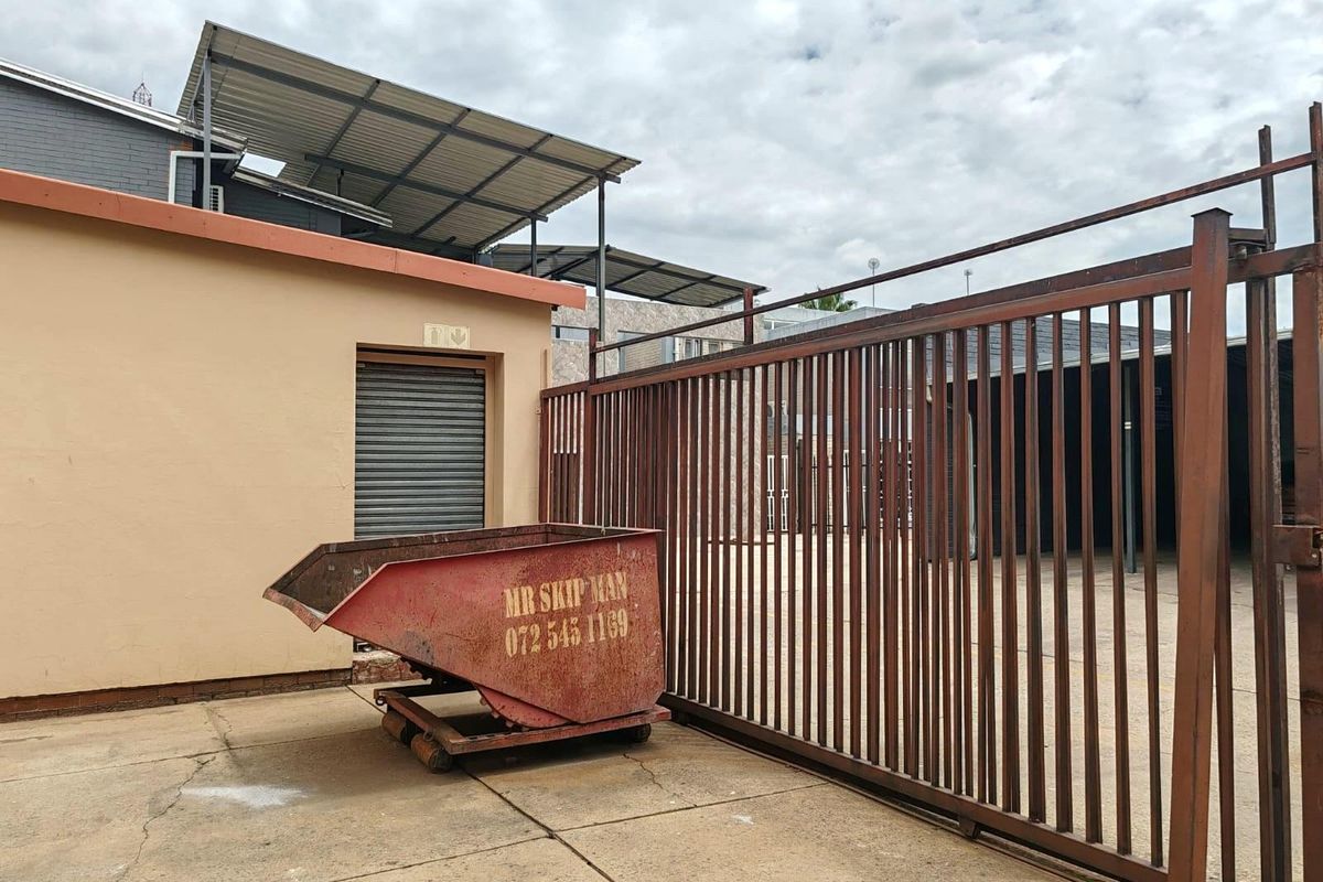 Exterior view of a commercial property featuring a large metal sliding gate, concrete paving, and a roller shutter door entrance.