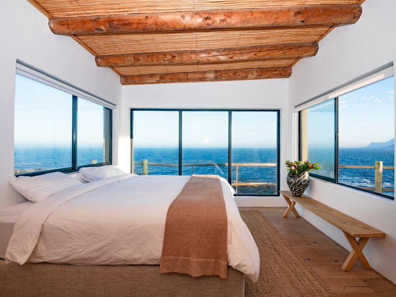 A bright bedroom featuring exposed timber beam ceilings, bamboo detailing, and expansive windows offering panoramic ocean views.