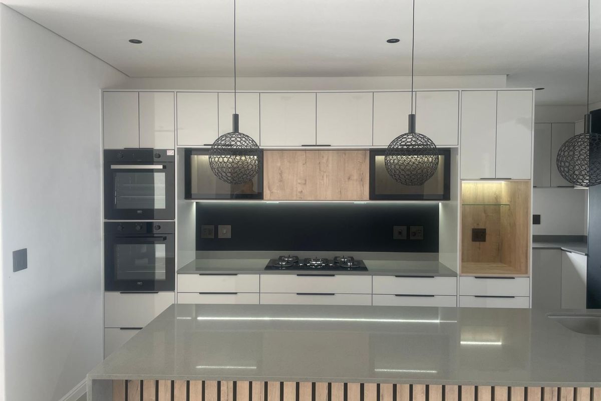 Modern kitchen featuring sleek white and wood-tone cabinetry, stone countertops, a central island, and built-in appliances with integrated lighting.