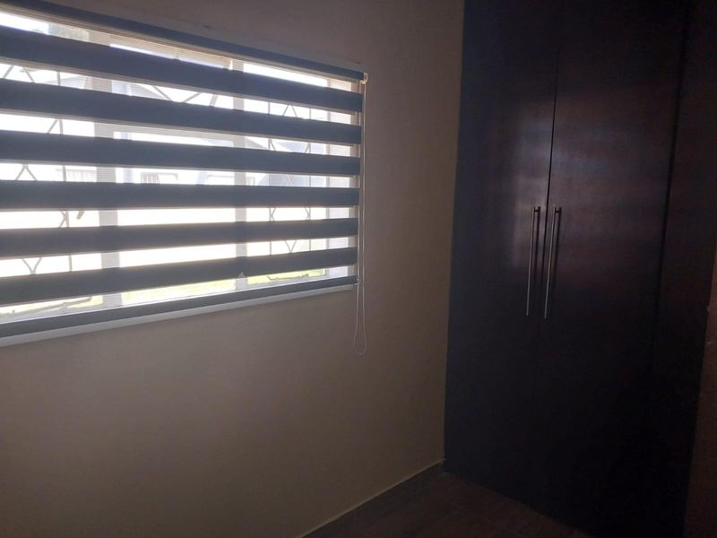 A clean, empty bedroom featuring a dark built-in wardrobe and a window fitted with modern roller blinds and security bars.