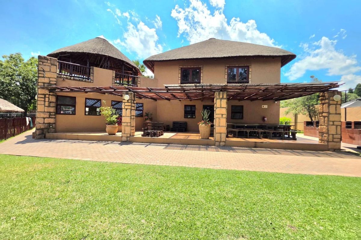Rear view of a two-story property featuring a thatched roof, stone-clad pillars, and a spacious paved patio area overlooking a well-maintained lawn.