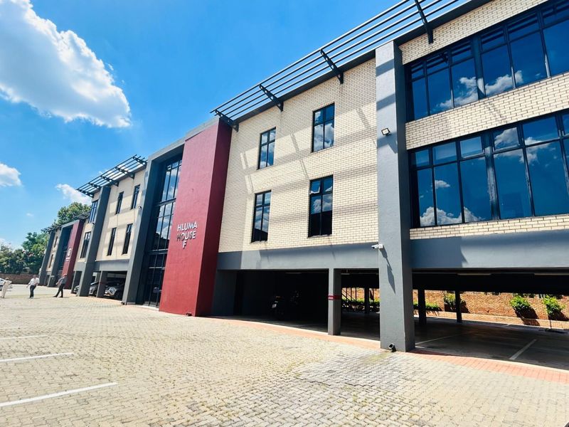 Modern commercial office building exterior featuring brick facade, large glass windows, and covered ground-level parking area.