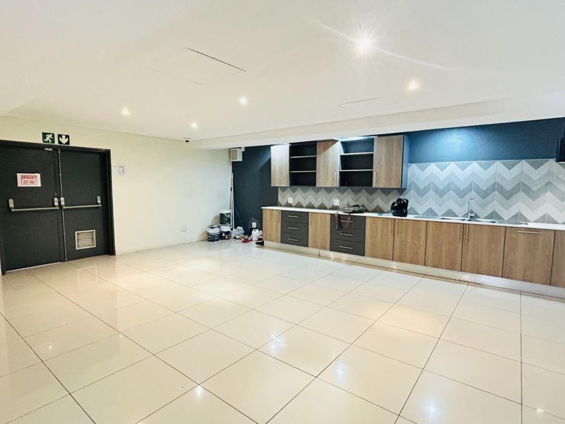 A spacious commercial kitchenette featuring modern wood-finish cabinetry, chevron-patterned backsplash, integrated sink, and tiled flooring with double emergency exit doors.