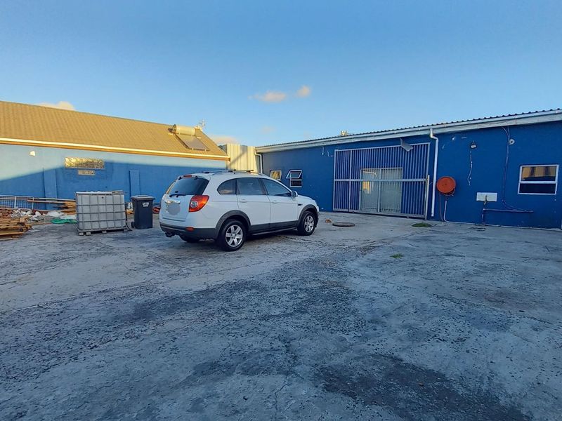 Rear exterior yard area featuring a concrete surface, metal sliding gate, and blue painted building facade with visible exterior plumbing and electrical fixtures.