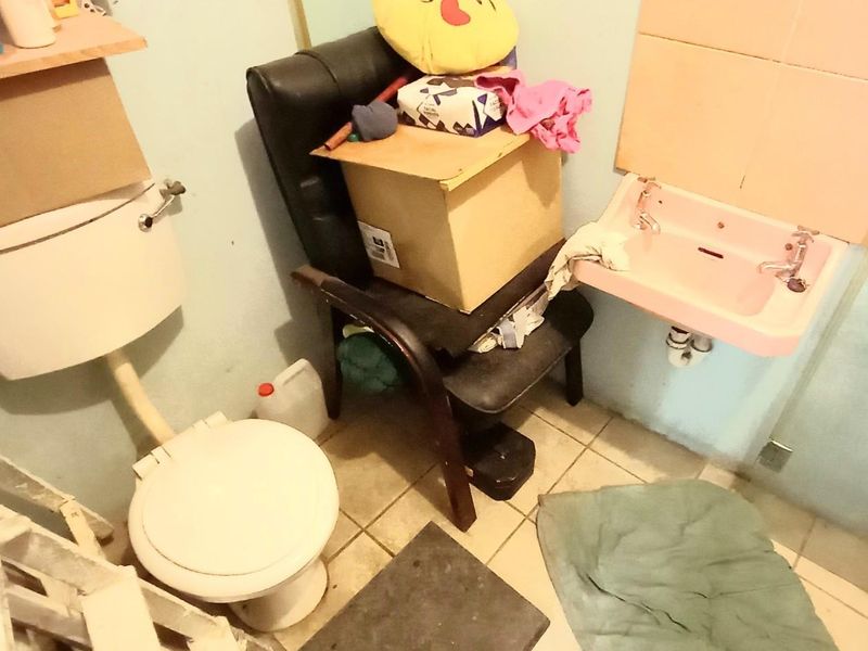 A small, dated bathroom featuring a wall-mounted basin and standard toilet, currently cluttered with personal items and in need of renovation.