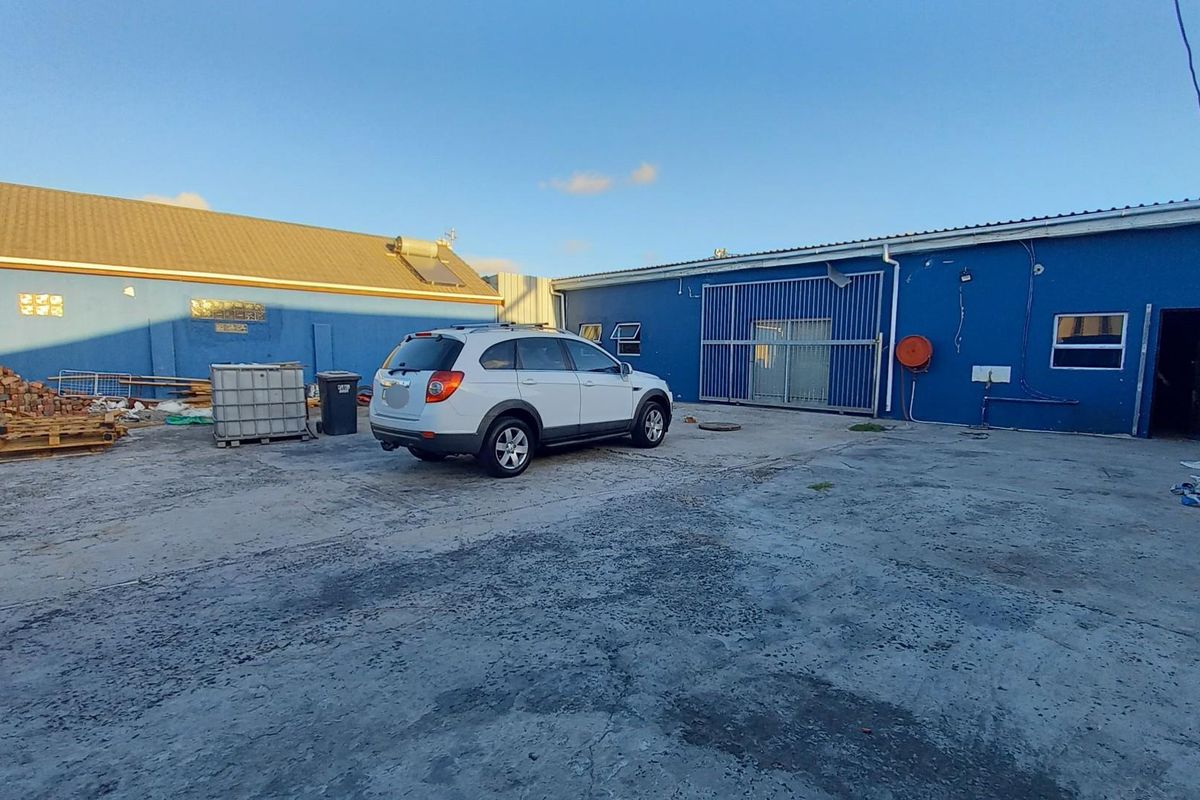 Rear exterior yard area featuring a concrete surface, metal sliding gate, and blue painted building facade with visible exterior plumbing and electrical fixtures.