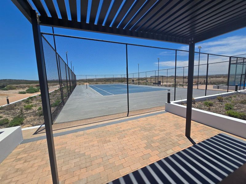 A well-maintained outdoor tennis court featuring perimeter fencing, floodlights, and an adjacent paved patio area with a modern pergola structure.