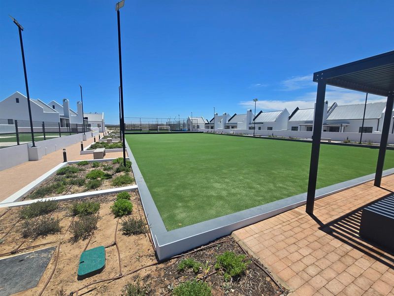 A well-maintained outdoor artificial turf bowling green surrounded by paved walkways, landscaped garden beds, and modern residential units.