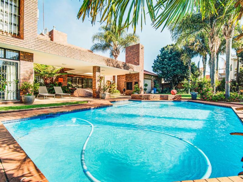 Spacious outdoor entertainment area featuring a large swimming pool, covered patio with built-in braai, and established garden surroundings.