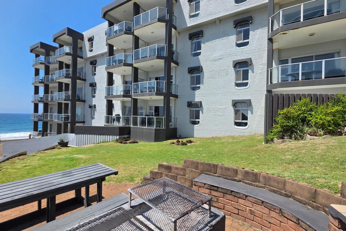 Exterior view of an apartment complex featuring private balconies, a communal lawn area, and a built-in brick braai facility overlooking the ocean.